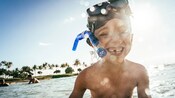A smiling young boy in shallow ocean water, his snorkeling mask worn high on his forehead 