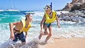 A boy and a girl in snorkel gear laughing as they emerge out of the water onto a seashore