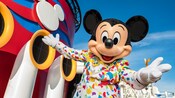 Mickey Mouse parado en la cubierta de un crucero, vestido con un traje adornado con papel picado.