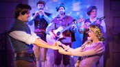 In a live show, Flynn Rider and Rapunzel hold hands and sing to each other as an acoustic band looks on