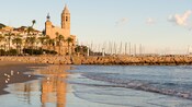 A cathedral stands next to the palm lined promenade and harbor in Stiges, Spain