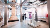 A large Celtic stone cross with biblical reliefs and 2 women's dresses on display in a museum setting