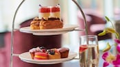 Uma taça de espumante perto de uma bandeja em camadas que contém pratos com macarons, scones e outras sobremesas em miniatura