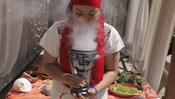 A young woman with a ceremonial cloth tied around her forehead inhaling a vapor wafting out of a large metal goblet