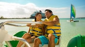 A man and woman steer an Aqua Cycle Water Trike with a Disney Cruise Line ship and a sailboat in the distance behind them