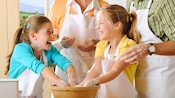 2 little girls in cooking aprons share a giggle as 2 adults look on