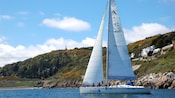 Passengers in a sailing yacht gliding past a rocky shoreline with a large home