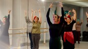 A dance instructor and her students facing a mirror as they perform a dance move with their hands in the air