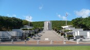 People stand in front of a memorial with a statue and stairs near trees and American flags