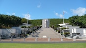 People stand in front of a memorial with a statue and stairs near trees and American flags