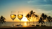Dos copas de vino sobre una mesa cerca de una playa tropical con palmeras