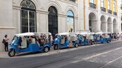 A line of 3-wheeled motorized eco tuk vehicles parked next to a historical building