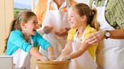 2 little girls in cooking aprons share a giggle as 2 adults look on