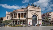 A stately, round, two-story building featuring a tall arch entrance, topped with horse statues