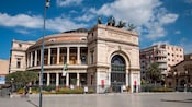 A stately, round, two-story building featuring a tall arch entrance, topped with horse statues