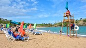 People lounging shoreside on a sandy beach while a lifeguard watches families playing in the water