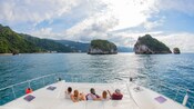 5 Guests sitting in the back of a catamaran looking out to a lush shoreline and rocky islets