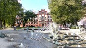 Árvores maduras e uma grande fonte com estátuas de bronze em frente a um edifício monumental