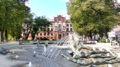 Árvores maduras e uma grande fonte com estátuas de bronze em frente a um edifício monumental