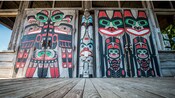 El exterior de madera de una cabaña de nativos americanos con un poste tótem y 2 murales que representan símbolos tótem dentro de ella