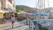A woman walking on a stone promenade lined with sidewalk cafes and docked boats