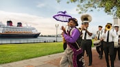 A man in cheery attire with a fluffy parasol leads a small band of musicians past a Disney cruise ship