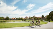 A male and female ride bikes past a small pond with a water fountain