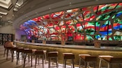 Stools in front of a long bar with a brilliant stained glass wall boasting many colors behind the bar