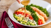 A cobb salad with avocado, bacon, lettuce, tomatoes, and more with a dressing being poured on from a hand above the plate