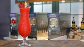 A frozen, blended cocktail in a Hurricane glass garnished with a strawberry and pineapple wedge sits on a counter in front of frozen drink machines and liquor bottles