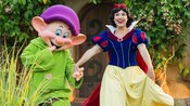 Near a quaint cottage doorway and greenery, a smiling Snow White walks hand in hand with Dopey, one of the 7 dwarfs