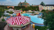 The pool area of Disney's BoardWalk Inn featuring 2 multistory buildings, leafy trees, a carousel shaped snack bar and a waterslide fashioned to look like a roller coaster