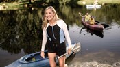  A woman holds an oar and walks out of a kayak at the side of a lake
