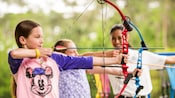 Two girls with bows and arrows take aim while an instructor looks on