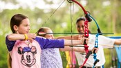 Dos niñas apuntan con arcos y flechas mientras un instructor observa