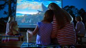 Una madre y una hija sentadas al aire libre se acurrucan mientras miran una proyección The Lion King en una pantalla