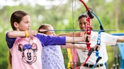 Dos niñas pequeñas apuntan con arcos y flechas mientras un instructor observa