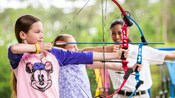 Deux petites filles visent des cibles avec des arcs et des flèches pendant qu’un instructeur les observe