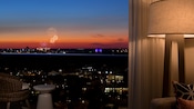 A hotel room window affords a view of fireworks exploding beyond a balcony at sunset