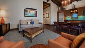 Una sala de estar con cadencia, una lámpara, un cuadro de pared, un sofá, 2 mesas ratonas, 2 sillas tapizadas y una cocina integrada con asientos en la encimera