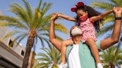 Una niña pequeña sentada en los hombros de su padre en un entorno al aire libre con palmeras Una niña pequeña sentada en los hombros de su padre en un entorno al aire libre con palmeras