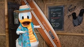 Donald Duck, dressed in a Hawaiian shirt and straw hat, posing in front of a Polynesian style structure beside a surfboard and a message board listing water conditions