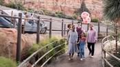 A male Cast Member holds Mickey balloons as a family of 4 walks up a trail in Radiator Springs 