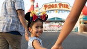 A smiling young girl, wearing Mickey ears with a bow, holds hands with two adults as she walks in front of Pixar Pier