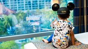 A kid wearing a Mickey ears hat sits on a bed and points toward a window inside the Disneyland Hotel A kid wearing a Mickey ears hat sits on a bed and points toward a window inside the Disneyland Hotel