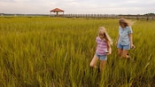 Mother and daughter trek through a field of marshy reeds, and in the background, a wooden walkway