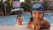 A little girl with a Mickey snorkle mask leans on the side of the pool while her dad watches