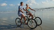 A man and a woman ride bicycles on the beach's wet sand