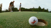 A Mickey golf ball teeters on a hole while a man and his wife admire his putt