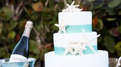 A wedding cake with seashell decorations next to a silver bucket with champagne